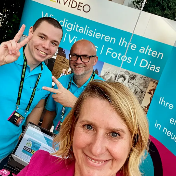 Das FORVIDEO Beratungsteam! Forvideo Beratungsteam mit einer Frau und 2 Männern die glücklich und zufrieden vor einer Forvideo Messewand stehen in deren Firmenfarben - aquablau und pink