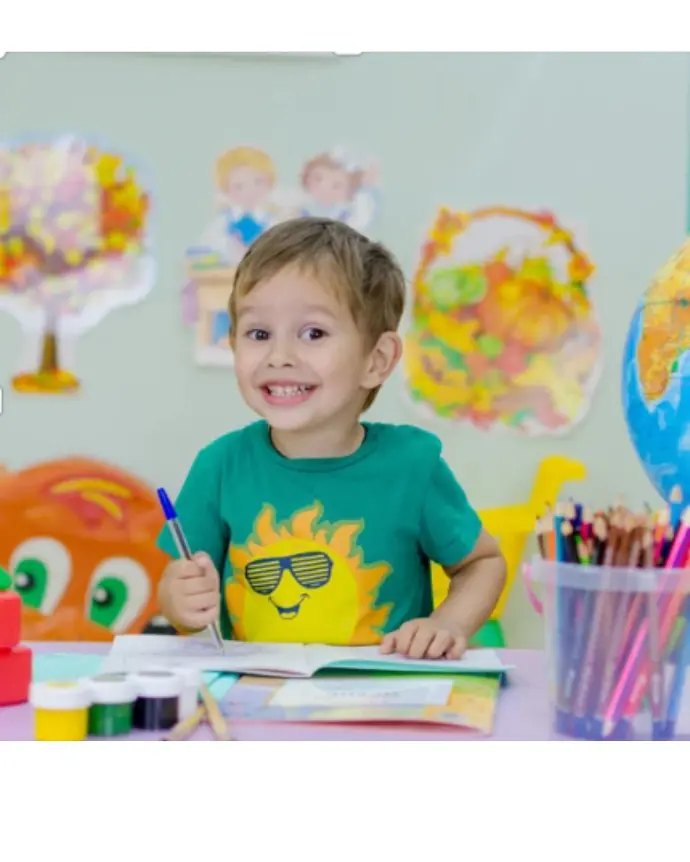 Ein Volksschuljunge sitzt voller Freude lächelnd bei seinem Maltisch und zeichnet mit vielen bunten Stiften ein Kinderkunstwerk 